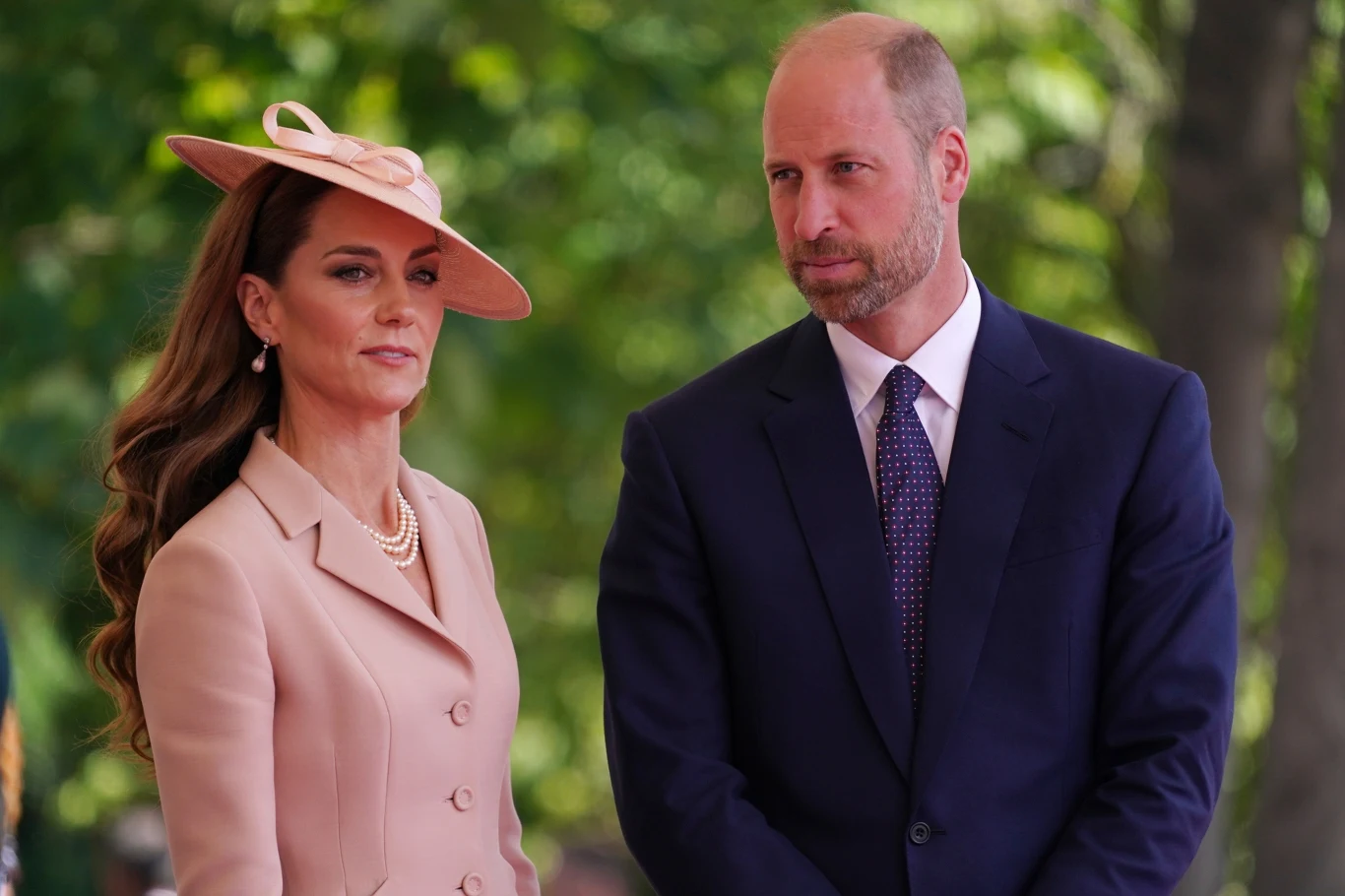 William nie ukrywa, że jest dumny ze swojej żony Elegancka kobieta w jasnoróżowym stroju i kapeluszu z falowanymi włosami obok mężczyzny w ciemnym garniturze i krawacie na tle zielonych drzew.