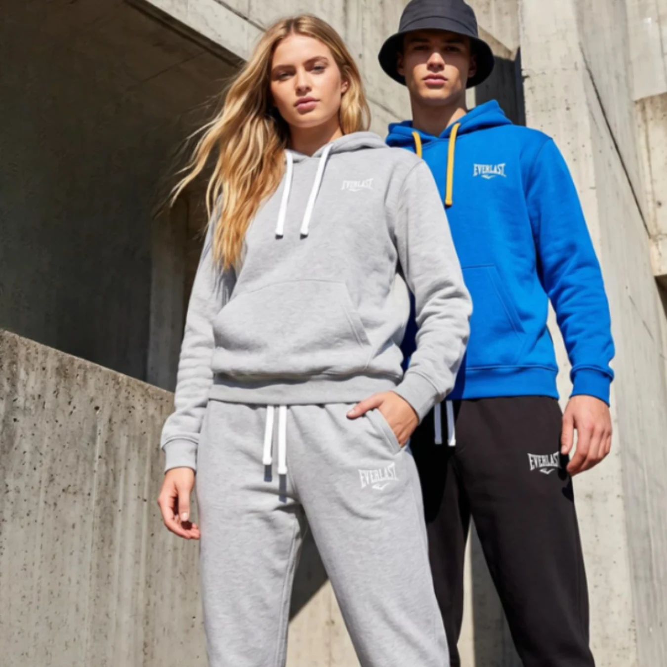 Everlast od 29,99 zł. Bluzy i dresy w supercenach Dwoje młodych osób w sportowych kompletach Everlast; kobieta w szarej bluzie i spodniach, mężczyzna w niebieskiej bluzie i czarnych spodniach oraz czarnym kapeluszu typu bucket stoją na tle betonowej architektury.
