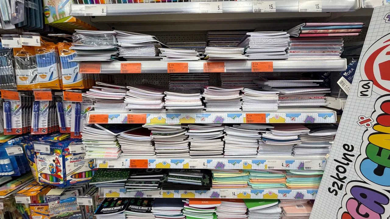 Zeszyty od 1 zł w Dealz. Do wyboru Herlitz, Oxford i Top 2000 Stosy różnorodnych zeszytów ułożone na półkach sklepowych, obok których znajdują się opakowania długopisów i kolorowych przyborów szkolnych.