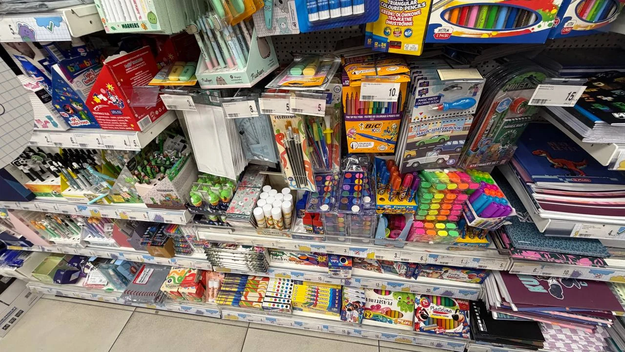Te drobiazgi też przydadzą się w piórniku Kolorowe przybory szkolne, w tym zestawy długopisów, flamastrów, kredek, plastelina, notesy, zeszyty i bloki, rozmieszczone na półkach w sklepie papierniczym.