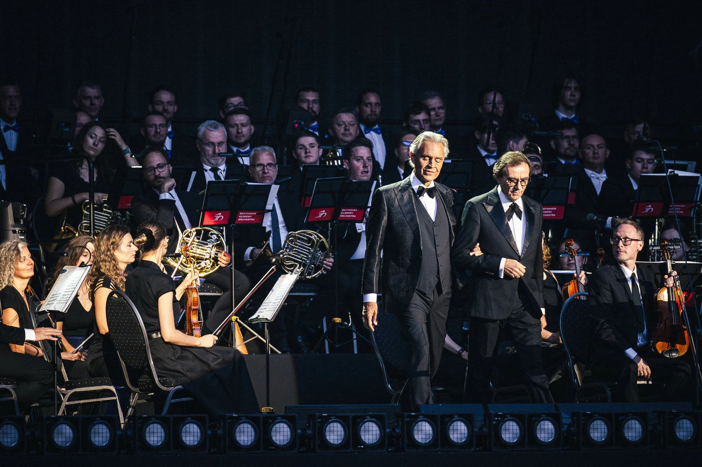 Gdańsk, Andrea Bocelli w Polsat Plus Arena podczas koncertu 14 sierpnia Dwóch elegancko ubranych mężczyzn w smokingu wychodzi na scenę przed orkiestrą symfoniczną, wśród muzyków z instrumentami smyczkowymi i nutami na pulpitach.
