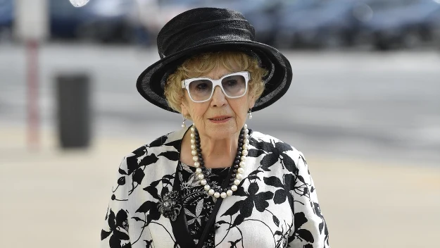 Starsza kobieta w eleganckiej czarno-białej marynarce w kwiatowy wzór, białych rękawiczkach i dużym czarnym kapeluszu, trzymająca bukiet różowych i fioletowych kwiatów oraz mająca naszyjnik z pereł i białe okulary w grubych oprawkach.