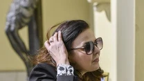Kobieta w ciemnych okularach przeciwsłonecznych i eleganckiej marynarce poprawia włosy, trzymając w ręku smartfona.