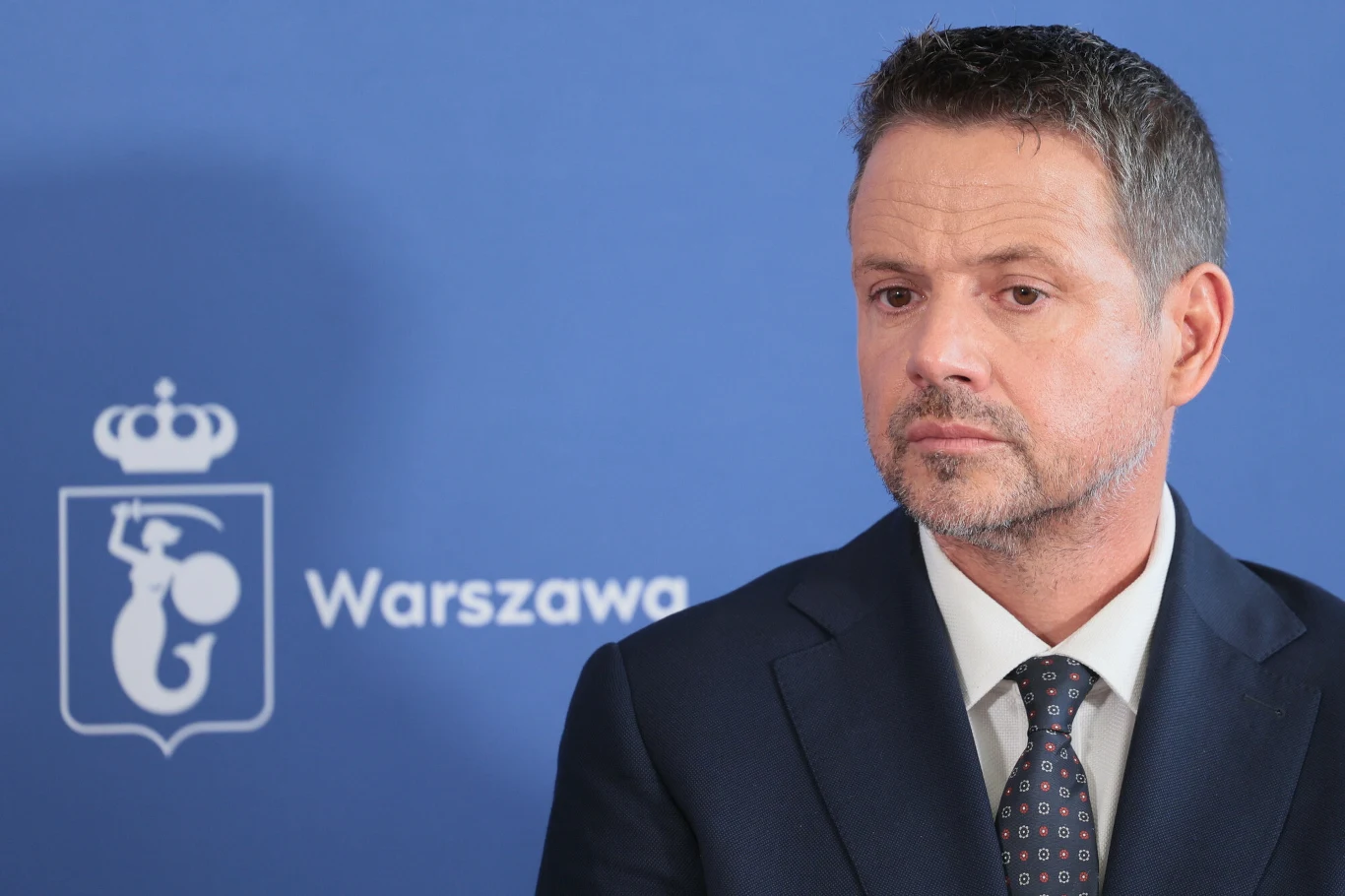 Prezydent Warszawy Rafał Trzaskowski Prezydent Warszawy Rafał Trzaskowski w garniturze i krawacie na tle niebieskiej planszy z herbem miasta Warszawy.