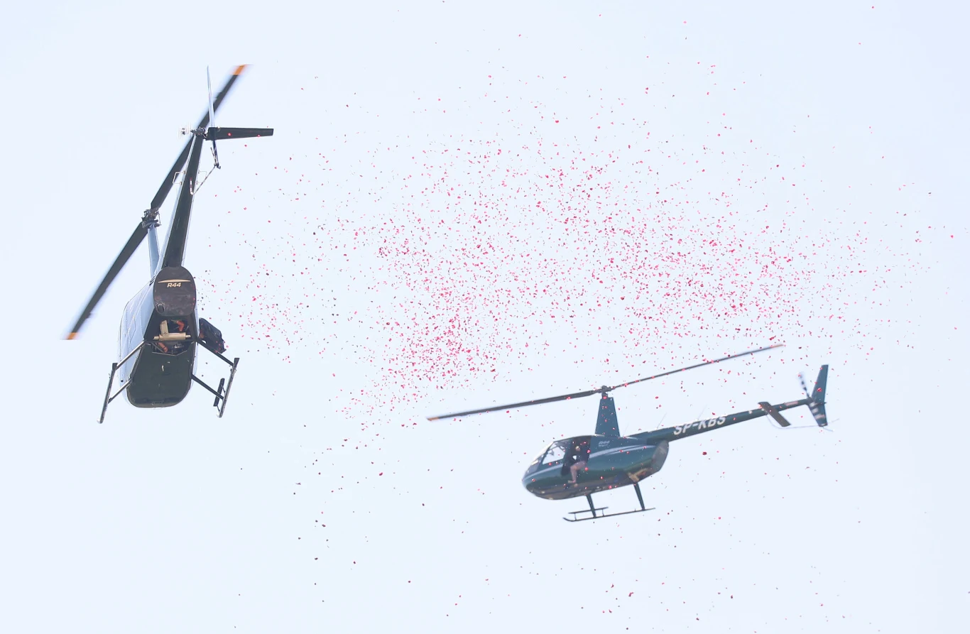 Dwa helikoptery unoszą się w powietrzu, a w ich otoczeniu unoszą się liczne czerwone konfetti rozsypujące się na jasnym tle nieba.
