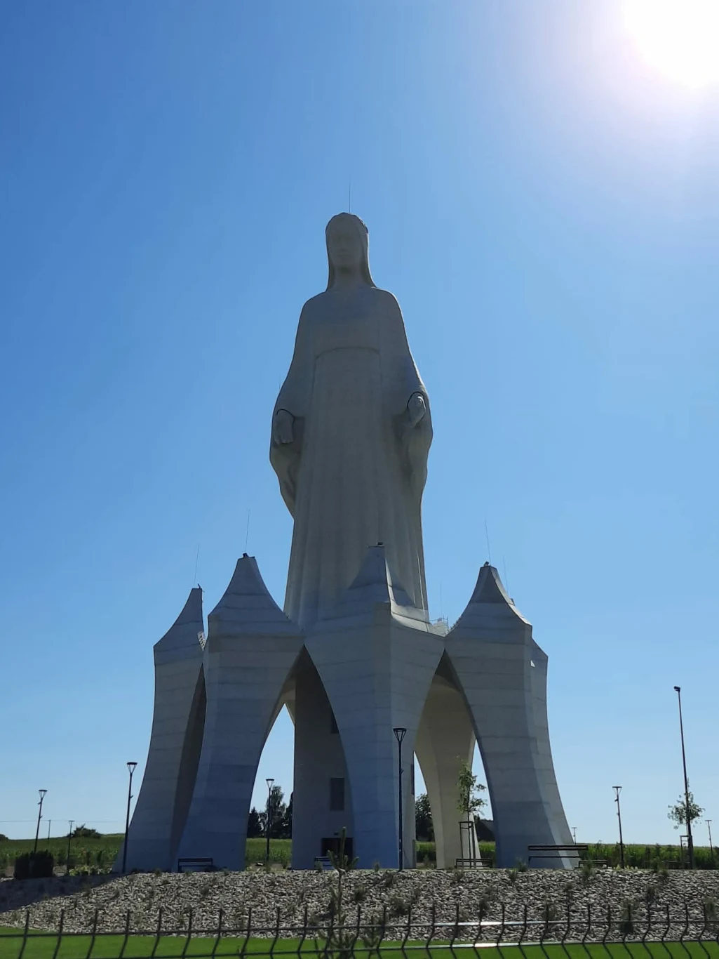 Monumentalna, jasna figura stojącej postaci na masywnej podstawie z wysokimi łukowatymi kolumnami, w otwartym, słonecznym terenie.