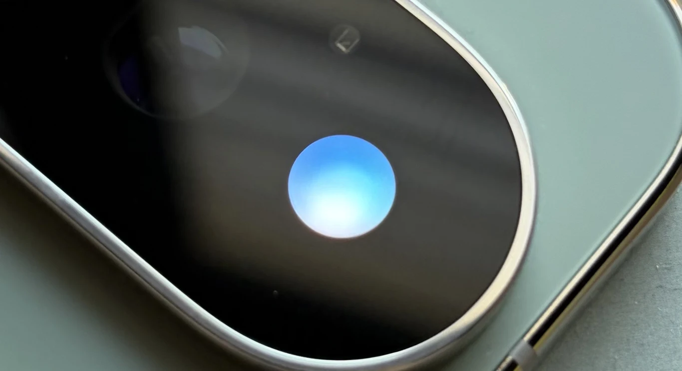 Funkcja HiLight z Google Pixel 11 Pro daje dostęp do powiadomień bez patrzenia na ekran. Obiektyw aparatu smartfona z odbijającym się niebieskim światłem, otoczony metalową ramką na tle zielonej obudowy urządzenia.