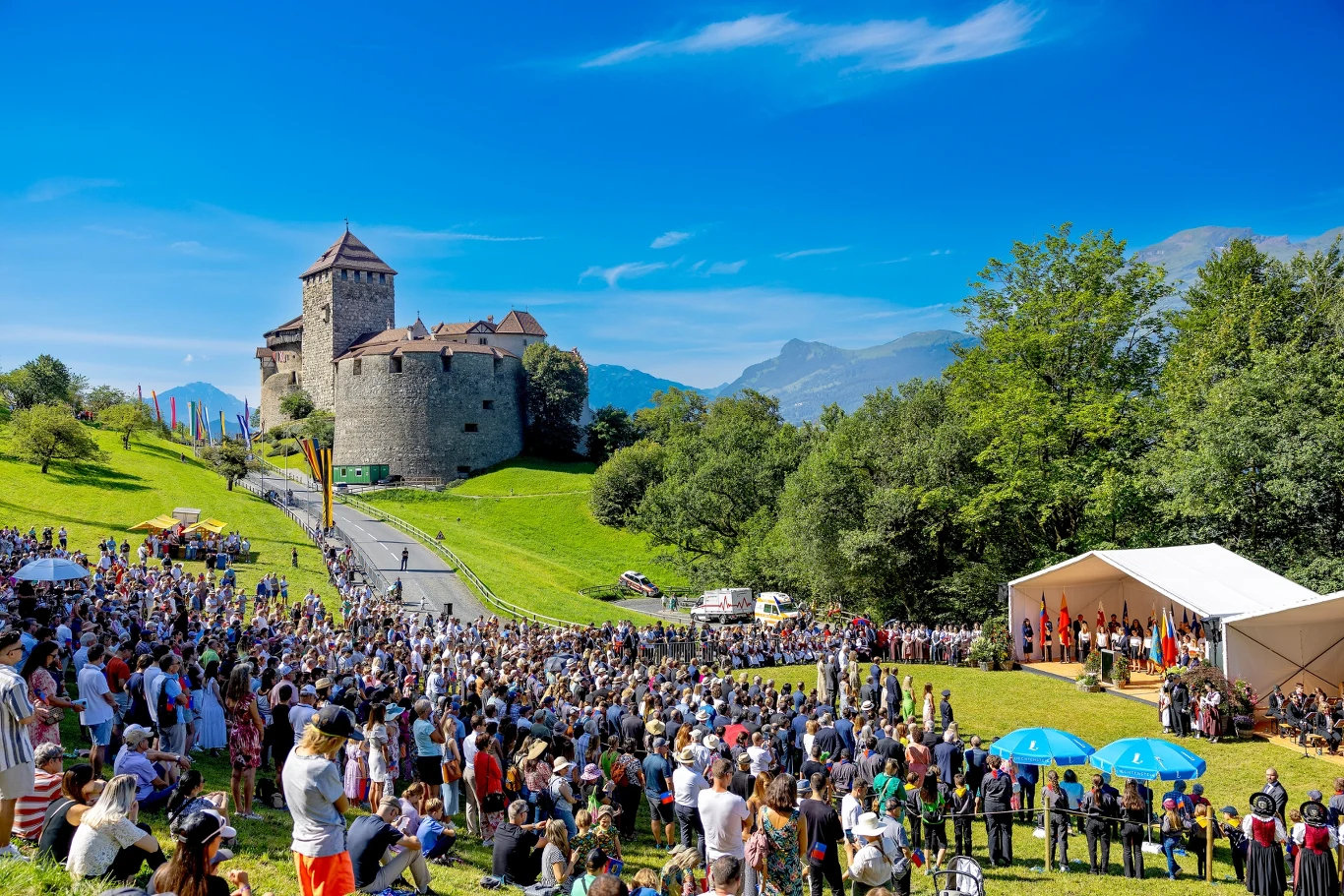 Obchody Święta Narodowego Liechtensteinu w Vaduz Tłum ludzi zgromadzony na zielonym wzgórzu przed historycznym zamkiem, obok rozstawionej sceny z flagami i osobami przemawiającymi, w tle widać góry pod błękitnym niebem.