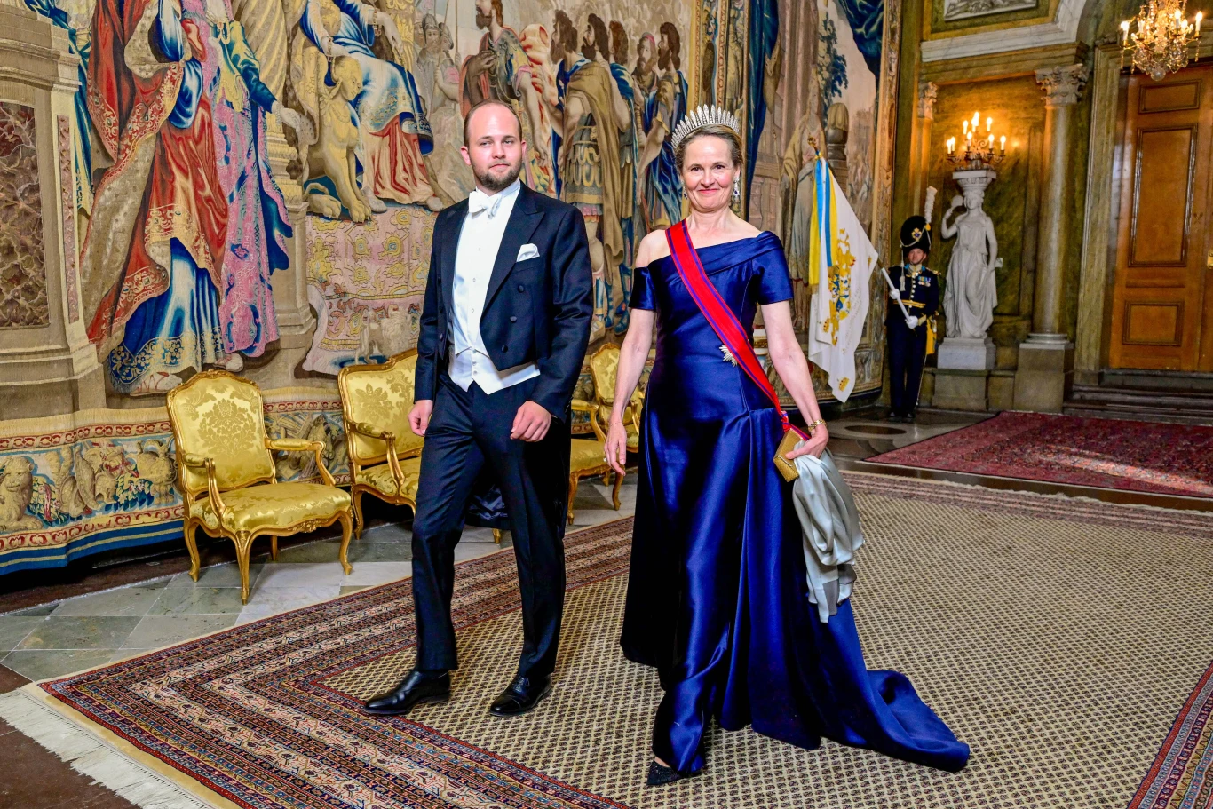 Drugi (po ojcu) w kolejce do tronu Liechtensteinu Józef Wacław z matką, księżną Zofią Elegancka kobieta w granatowej sukni z tiarą i szarfą oraz mężczyzna w smokingu kroczą przez bogato zdobioną salę z gobelinami i złotymi fotelami.