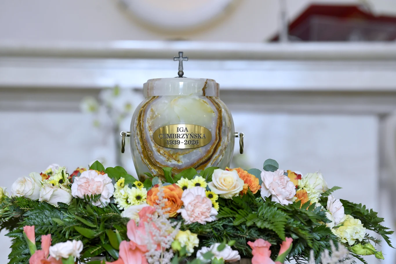 Pogrzeb Igi Cembrzyńskiej marmurowa urna z tabliczką imienną, ustawiona pośród bukietów świeżych kwiatów w jasnych kolorach