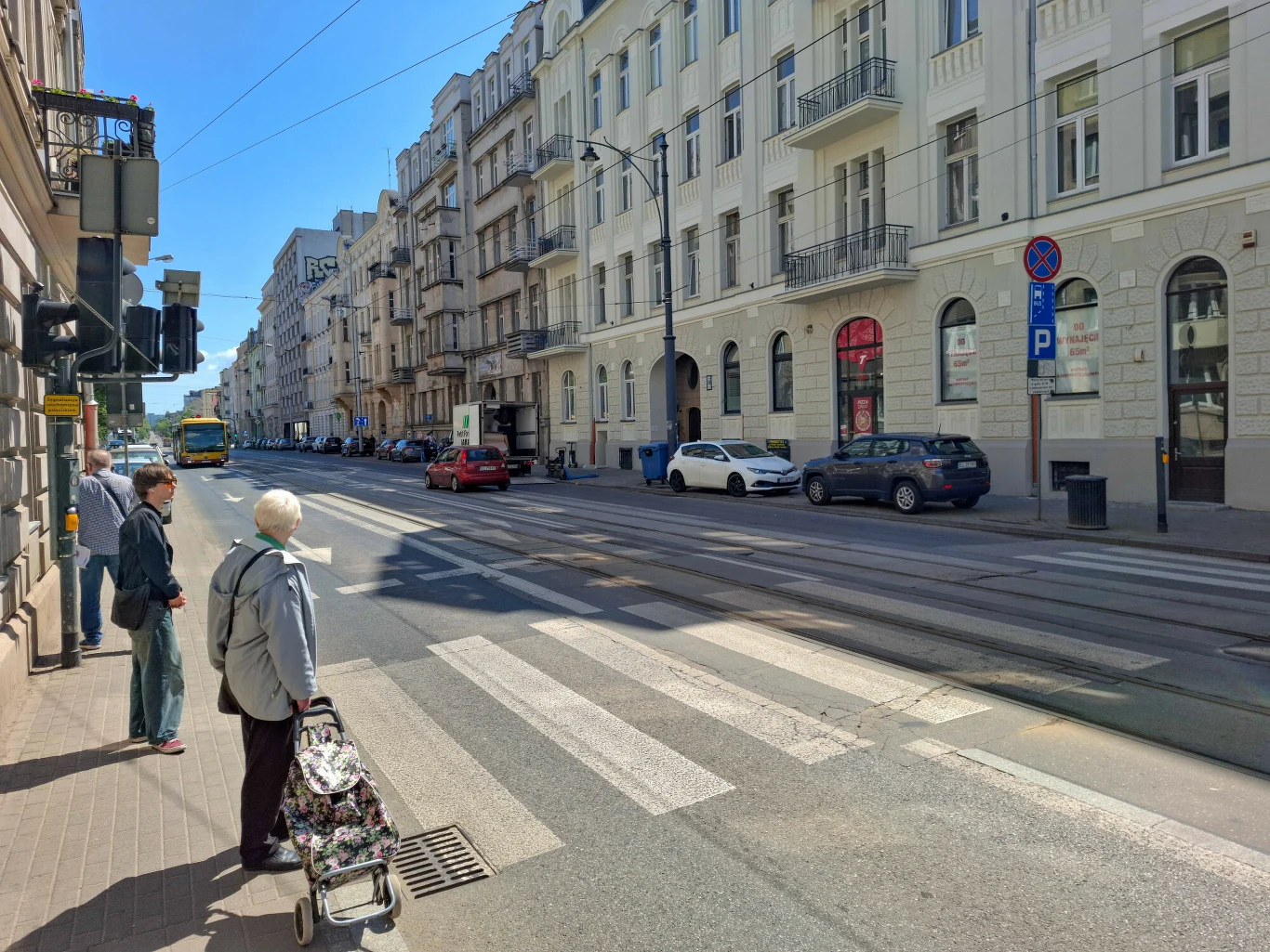 Fakt, że w Polsce piesi czekają na czerwonym świetle nawet, gdy nic nie jedzie, zadziwił Włocha Dwie osoby czekające przy przejściu dla pieszych na tle miejskiej ulicy z zaparkowanymi samochodami i wysokimi, jasnymi kamienicami po obu stronach jezdni.