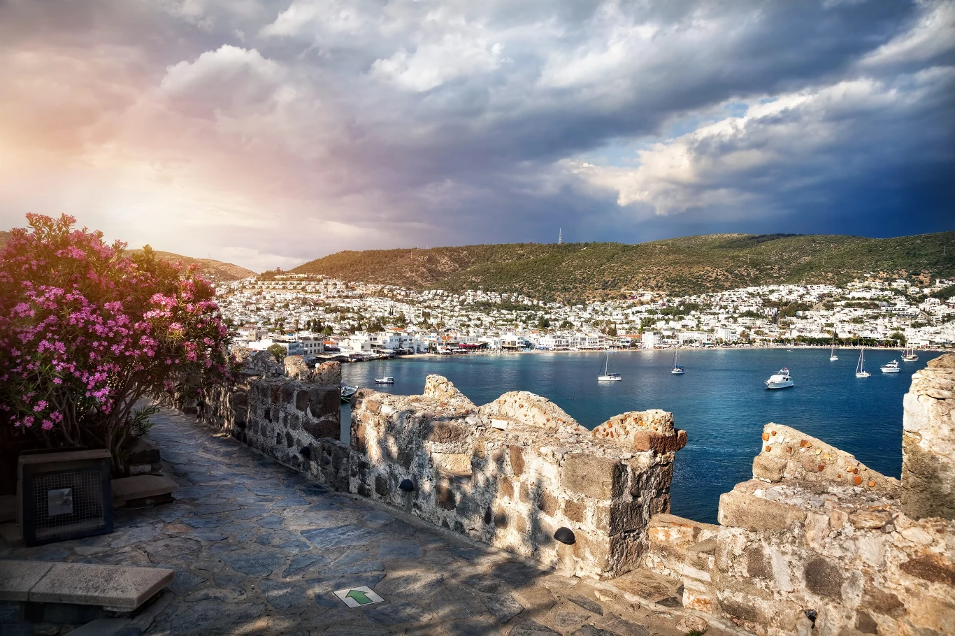 Bodrum zachwyca Zabytkowy kamienny mur na pierwszym planie, kolorowe krzewy kwitnącej bugenwilli po lewej stronie oraz rozległa zatoka z cumującymi jachtami i jasną zabudową miasteczka na tle zielonych wzgórz pod pokrytym chmurami niebem.