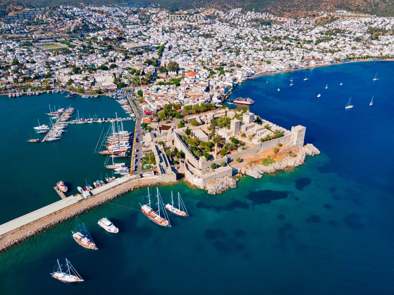 Widok na zamek i marinę w Bodrum Kamienna forteca położona na skalistym cyplu otoczona niebieskim morzem i mariną z cumującymi żaglówkami, w tle rozległe, jasne zabudowania miasta.