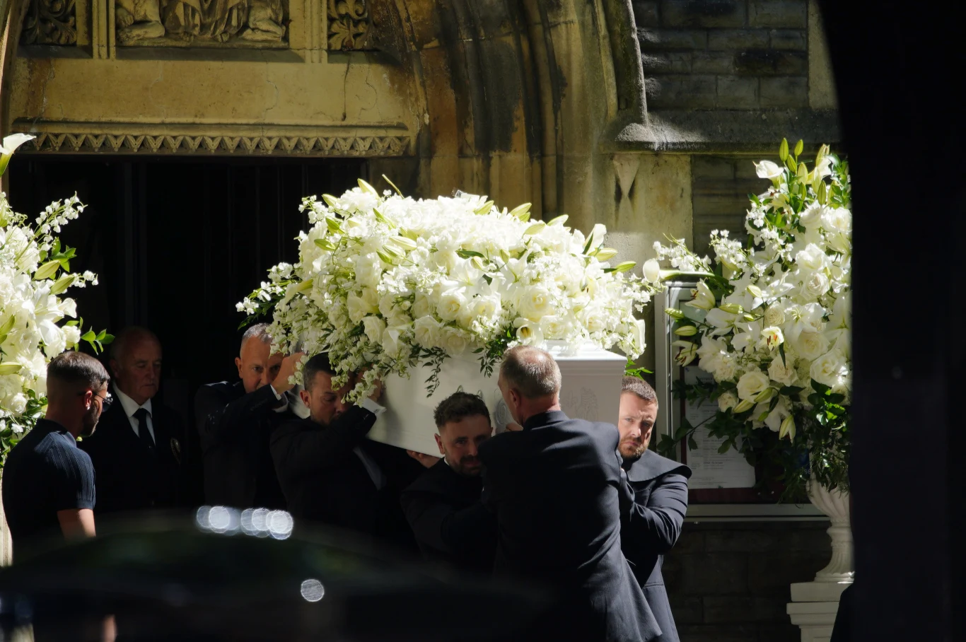 Kilku mężczyzn niesie białą trumnę ozdobioną obfitymi bukietami białych kwiatów, w otoczeniu bogato udekorowanego wejścia do budynku.
