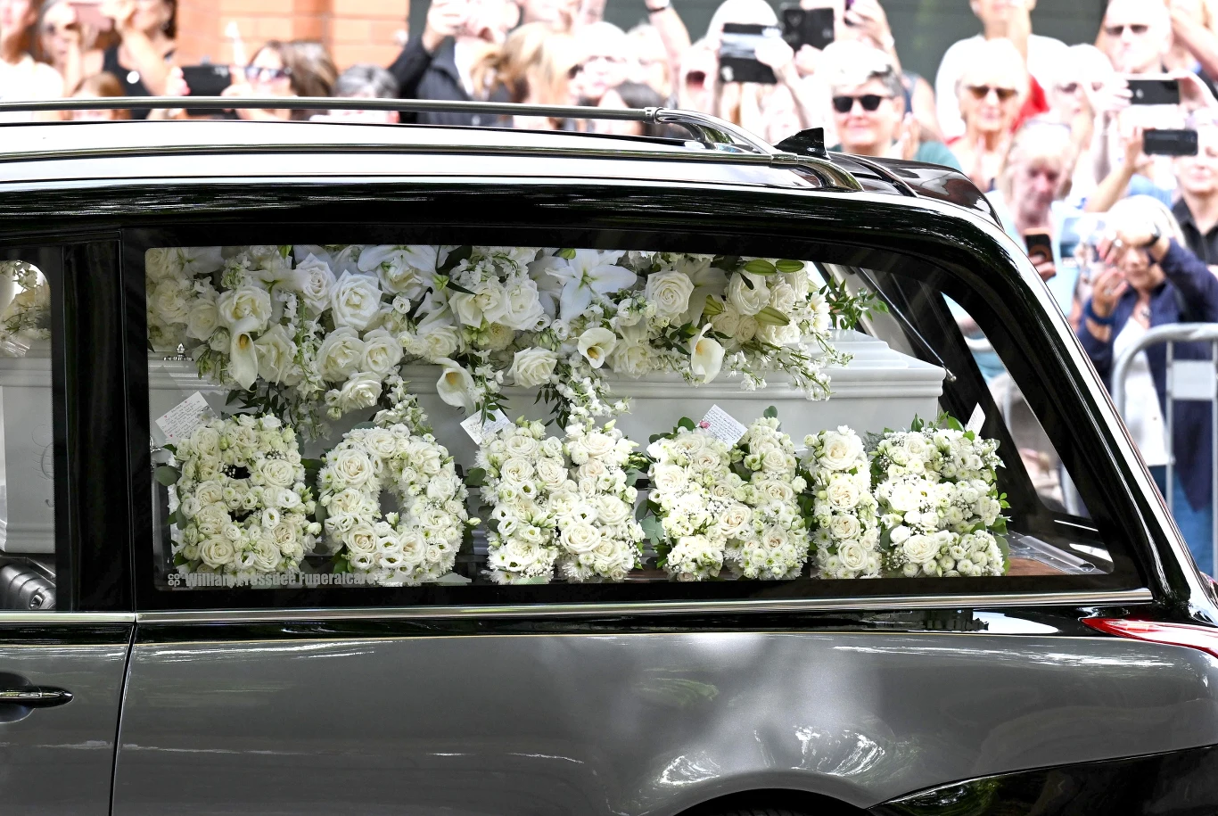 Biały karawan z trumną ozdobioną białymi kwiatami, przy której umieszczony jest okazały kwiatowy napis utworzony z białych róż, otoczony przez tłum żegnających osób fotografujących procesję.