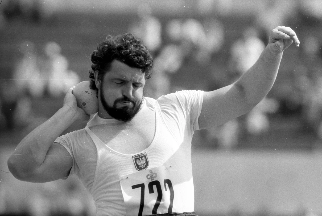 Władysław Komar Kulomiot w trakcie przygotowania do wykonania pchnięcia kulą, ubrany w sportową koszulkę z godłem i numerem startowym 722, skupiony na wykonaniu technicznego ruchu.
