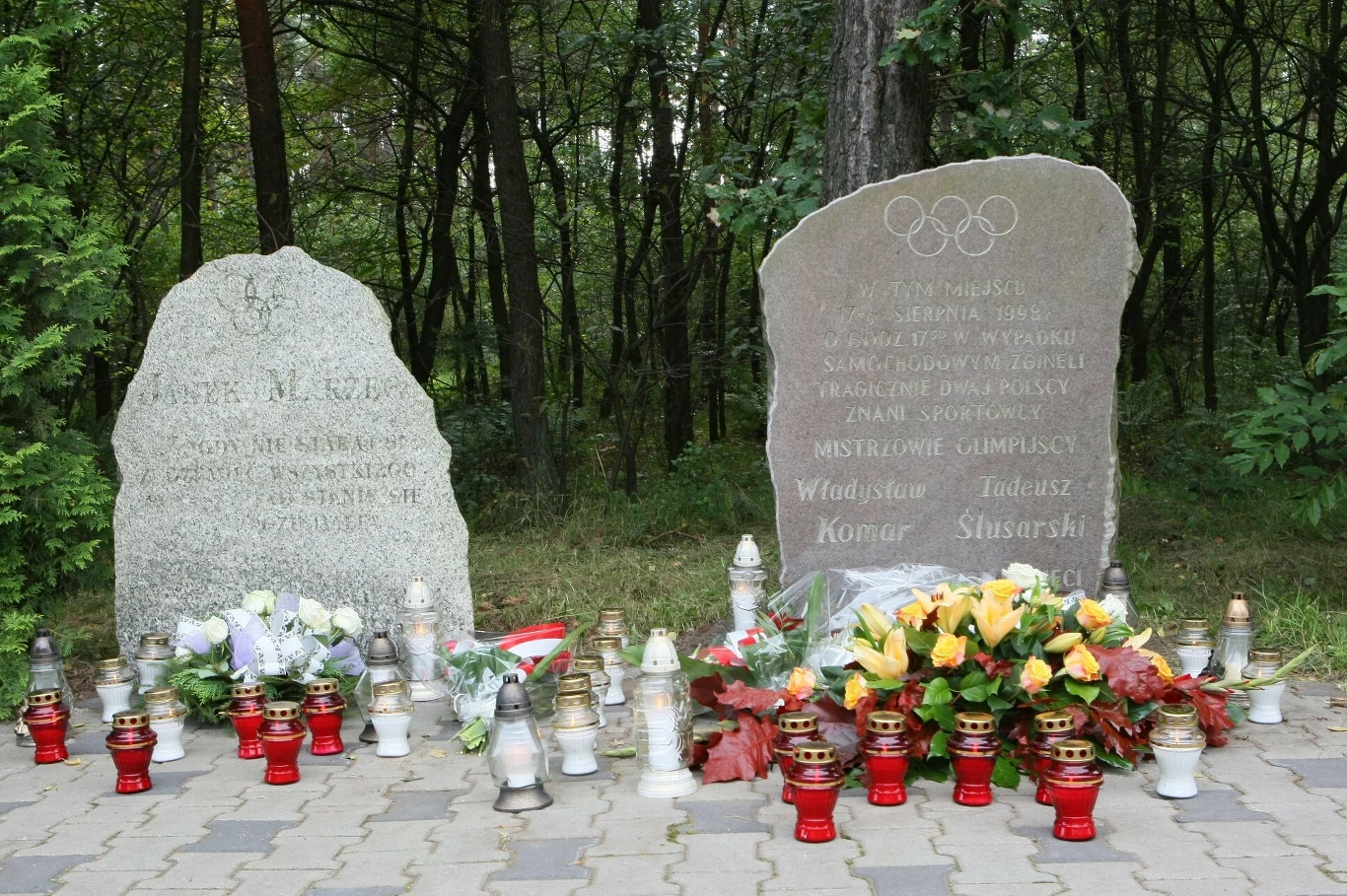 Tablice pamiątkowe na miejscu tragedii Dwa sąsiadujące nagrobki z licznymi zniczami i kwiatami ustawionymi przed nimi, pośród drzew i zieleni, z widocznymi napisami upamiętniającymi sportowców olimpijskich.
