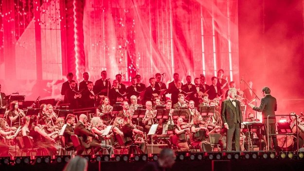 Orkiestra symfoniczna i chór wykonujące utwór na scenie z efektownym czerwonym oświetleniem, z dyrygentem i solistą na pierwszym planie.