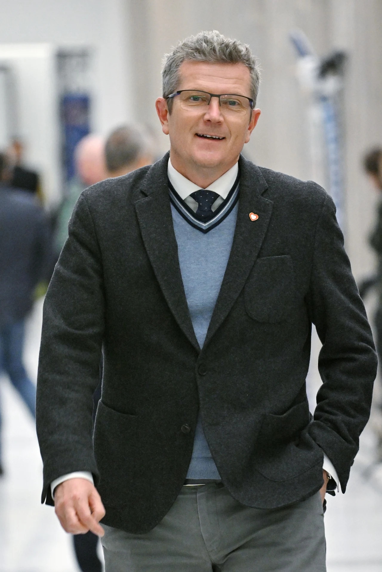Marcin Bosacki, wiceminister spraw zagranicznych Mężczyzna w szarym garniturze, niebieskim swetrze i okularach idzie korytarzem z ręką w kieszeni, uśmiechnięty, z krótkimi siwymi włosami.