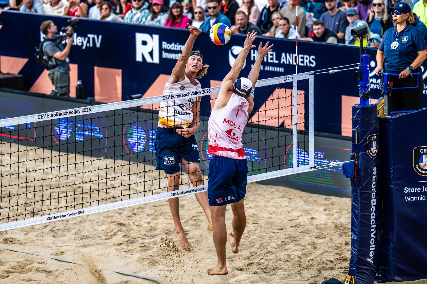 Dwóch siatkarzy rywalizuje przy siatce podczas meczu siatkówki plażowej, jeden z nich uderza piłkę, a drugi próbuje ją zablokować, wokół boiska zgromadzona publiczność kibicuje zawodnikom.