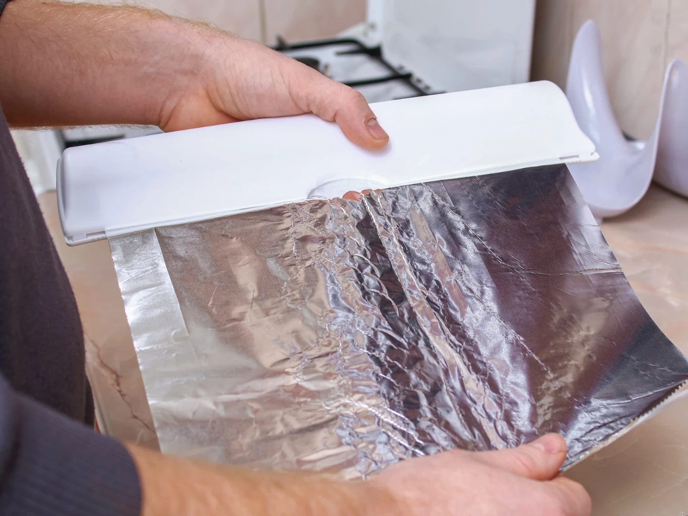 Ręce trzymają dozownik z folią aluminiową, która jest właśnie odcinana w kuchni na blacie obok kuchenki gazowej.