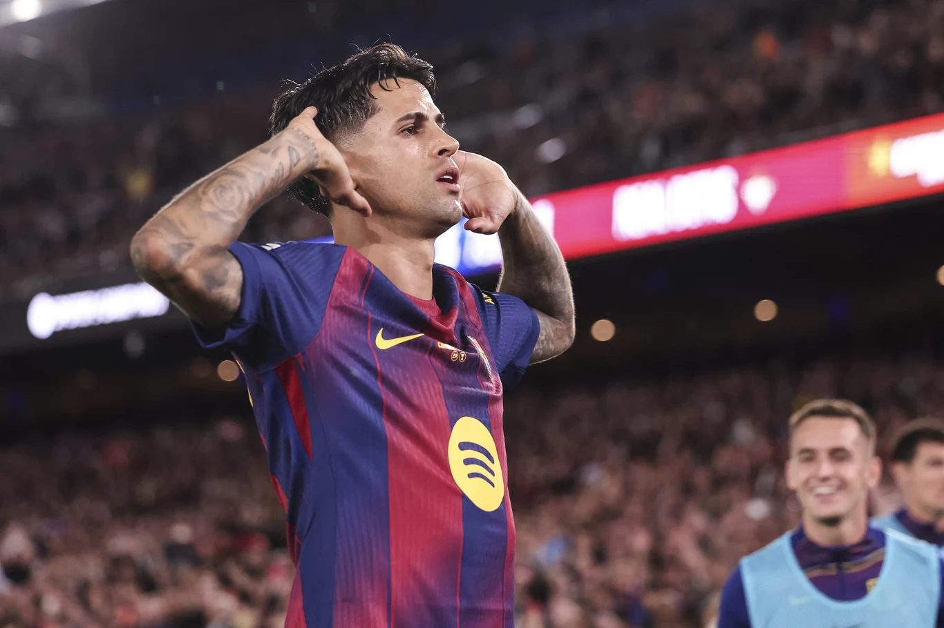 Piłkarz w koszulce FC Barcelony świętuje zdobycie gola, przykładając palce do uszu, w tle rozmazany tłum kibiców na stadionie.