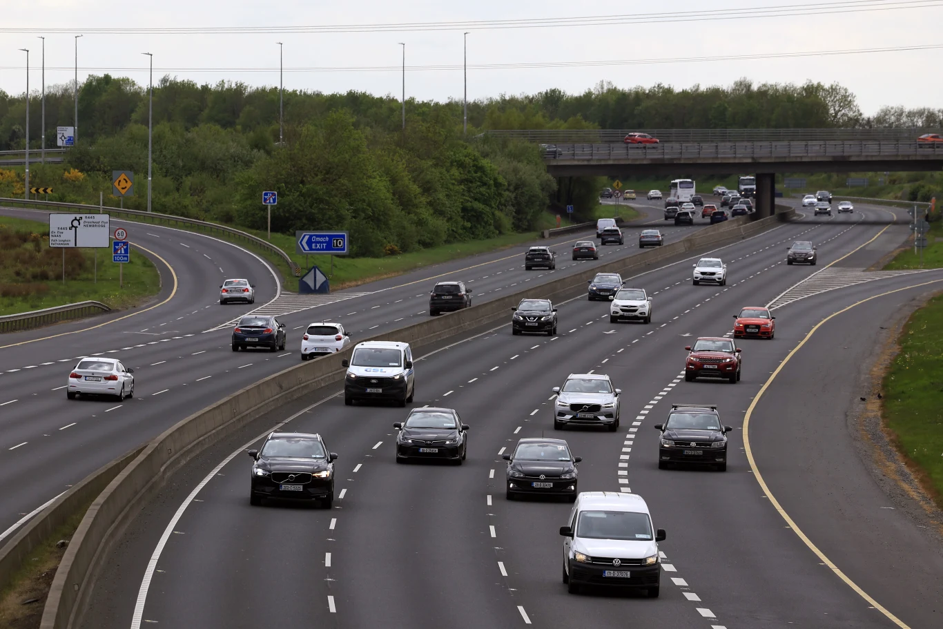 Ruchliwa autostrada z wieloma pasami w obu kierunkach, samochody osobowe i dostawcze poruszające się pod wiaduktem, otoczone pasem zieleni i zaroślami.