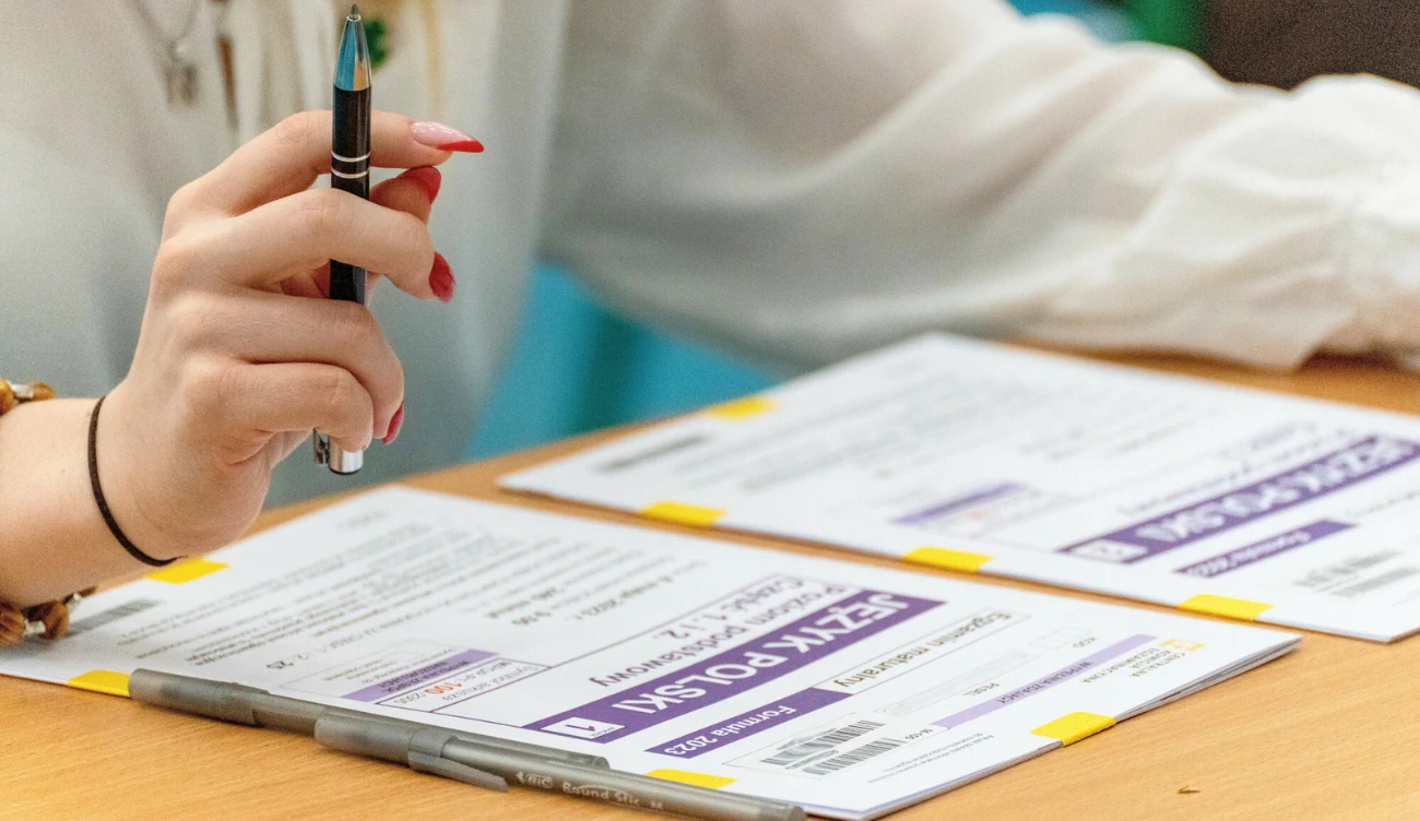 Matura poprawkowa 2026 z języka polskiego Dłoń z długopisem nad arkuszami egzaminacyjnymi z języka polskiego, na stole dwa szare długopisy.