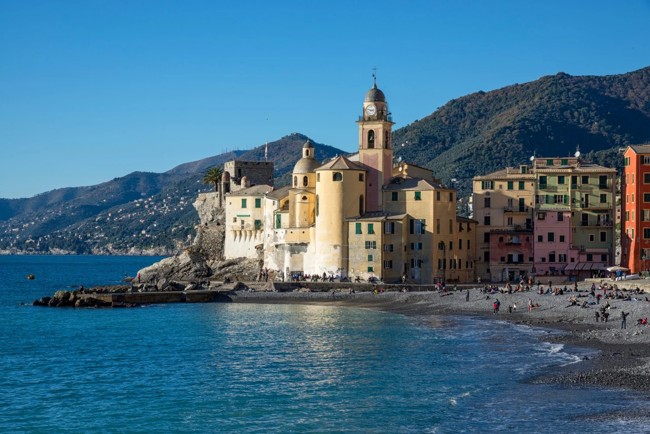 Camogli zamiast Portofino we wrześniu. Tu Liguria ma spokojniejsze oblicze Kolorowe budynki stojące tuż przy brzegu morza, w tym charakterystyczny kościół z wieżą, a na kamienistej plaży oraz wzdłuż promenady spacerują ludzie z widokiem na górzyste tło.