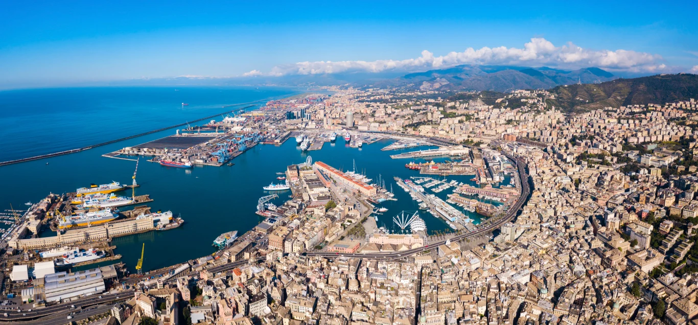 Genua Rozległy port morski z licznymi statkami oraz zwartą zabudową miejską otaczającą zatokę, w tle pasmo zielonych wzgórz i wybrzeże.