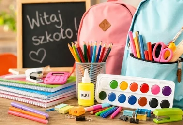 Przybory szkolne na biurku: zeszyty, kredki, farby i dwa plecaki przy tablicy z napisem Witaj szkoło