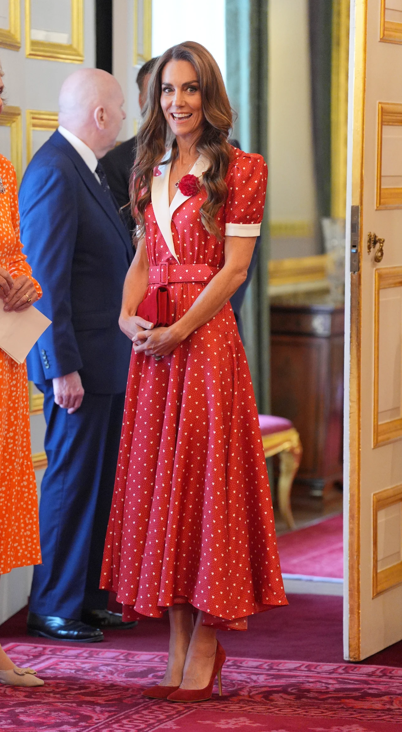Uśmiechnięta kobieta w długiej czerwonej sukience w białe kropki stoi na czerwonym dywanie w eleganckim wnętrzu, obok osób ubranych formalnie.