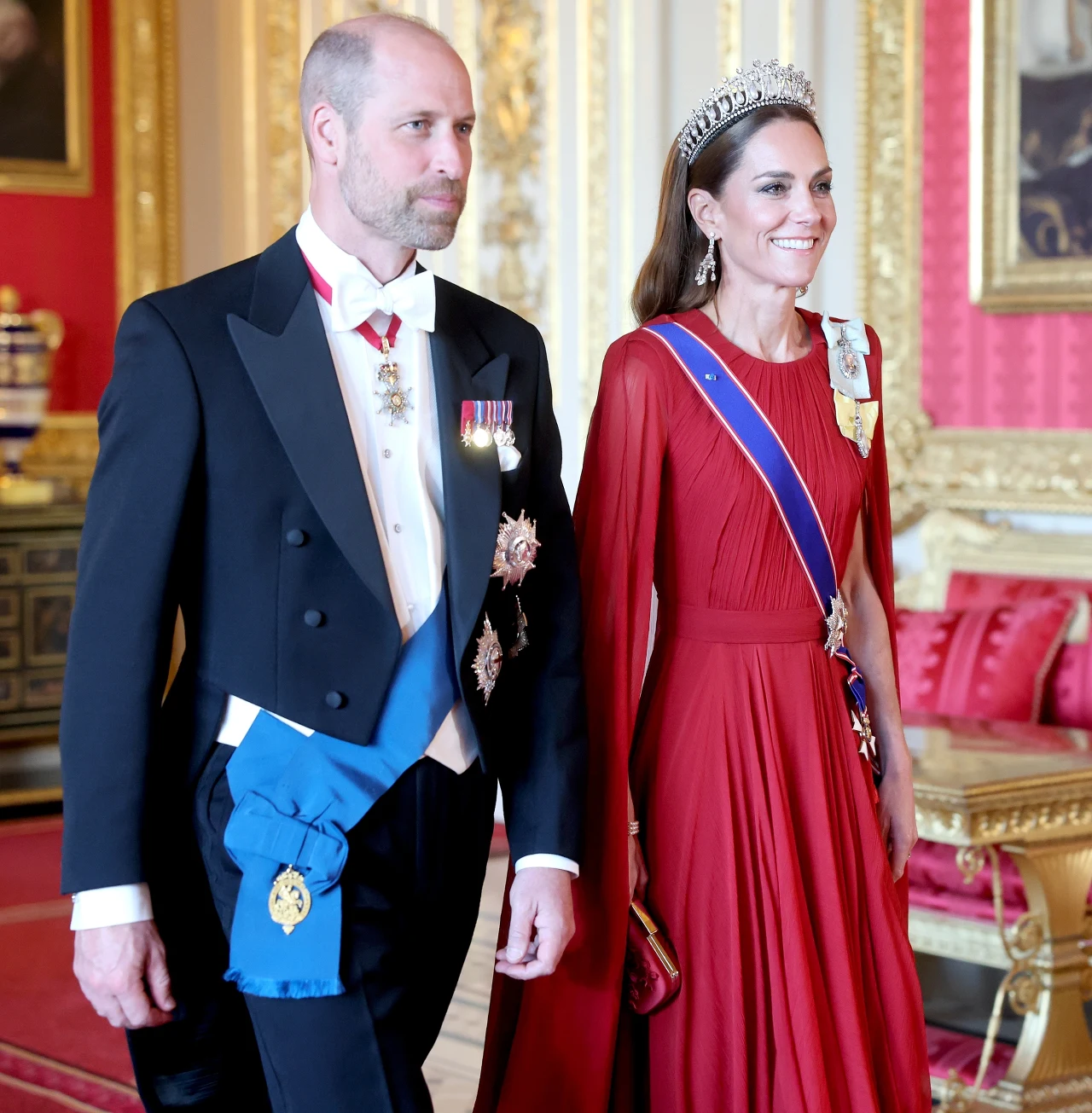 Elegancka para w bogato zdobionym wnętrzu pałacu, mężczyzna w smokingowym fraku z odznaczeniami i kobietą w czerwonej, długiej sukni, z tiarą oraz szarfą na ramieniu.