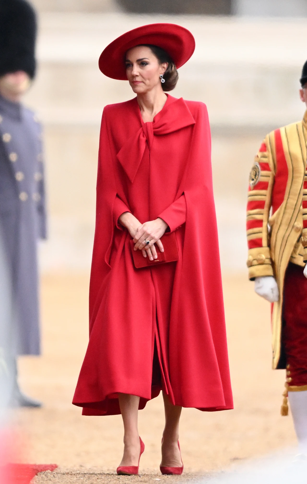 Elegancka kobieta w długim czerwonym płaszczu i szerokim kapeluszu w tym samym kolorze, trzymająca kopertówkę, stoi na zewnątrz w otoczeniu osób w historycznych strojach ceremonialnych.