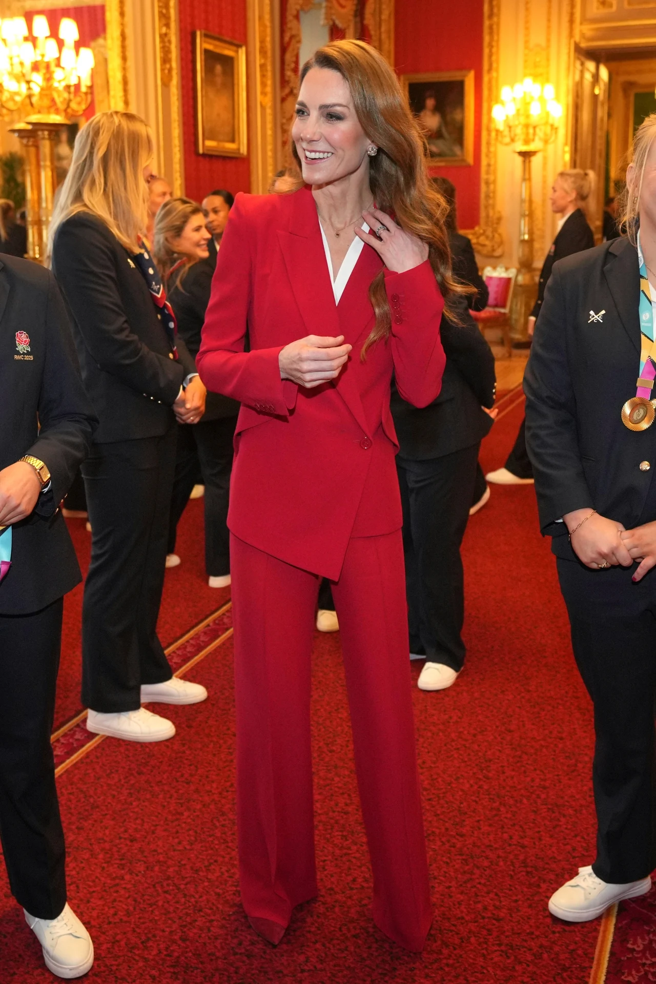 Kobieta w eleganckim czerwonym garniturze otoczona kilkoma osobami w ciemnych strojach, w tle złote oświetlenie i bogato zdobione ściany pałacowego wnętrza.