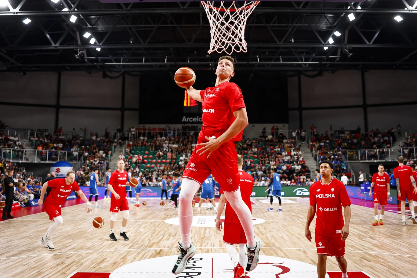 Igor Milicić Jr. Koszykarz w czerwonym stroju polskiej reprezentacji wykonuje wsad do kosza podczas rozgrzewki, otoczony przez innych zawodników swojej drużyny na tle pełnych trybun.