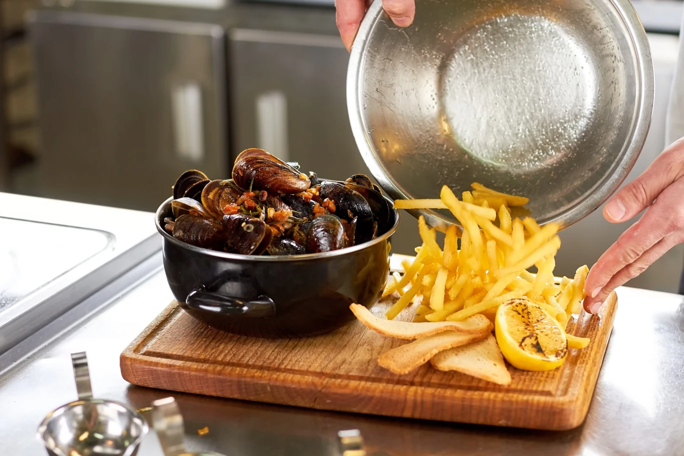 Duszone mule podawane z frytkami to rarytas kuchni belgijskiej i francuskiej Porcja muli w czarnym naczyniu, frytki wysypane na deskę do krojenia obok chrupiącego tosta i połówki grillowanej cytryny.