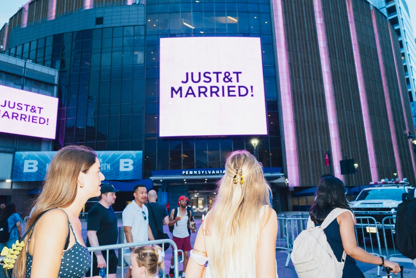 Trzy młode kobiety przechodzą obok wejścia do dużego budynku z neonowym napisem „JUST&T MARRIED!” na jasnym ekranie, wokół zgromadzeni są również inni piesi.