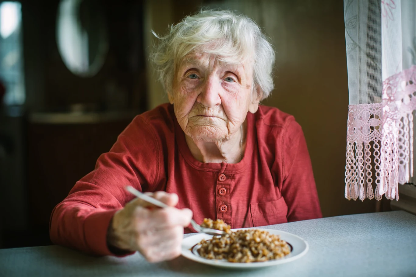 Seniorzy powinni jeść częściej, a mniej, żeby zahamować podjadanie rujnujące jelita Starsza kobieta w czerwonej bluzce je ciepły posiłek z kaszą przy stole obok okna z firanką.