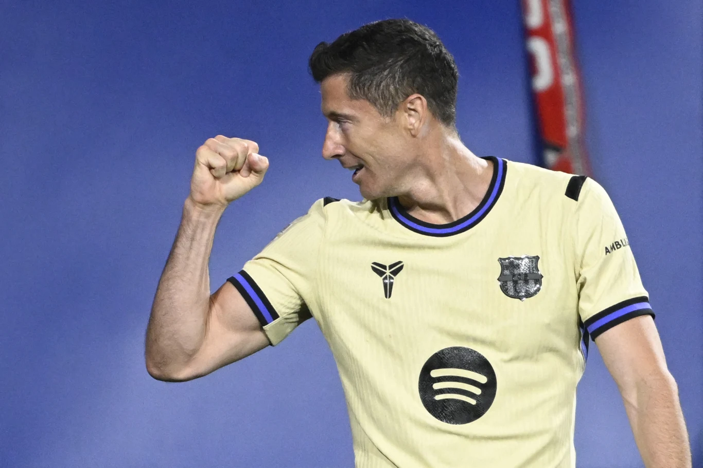 Robert Lewandowski w barwach FC Barcelona Robert Lewandowski w kremowej koszulce FC Barcelona z logo Spotify, na niebieskim boisku, zaciska pięść.