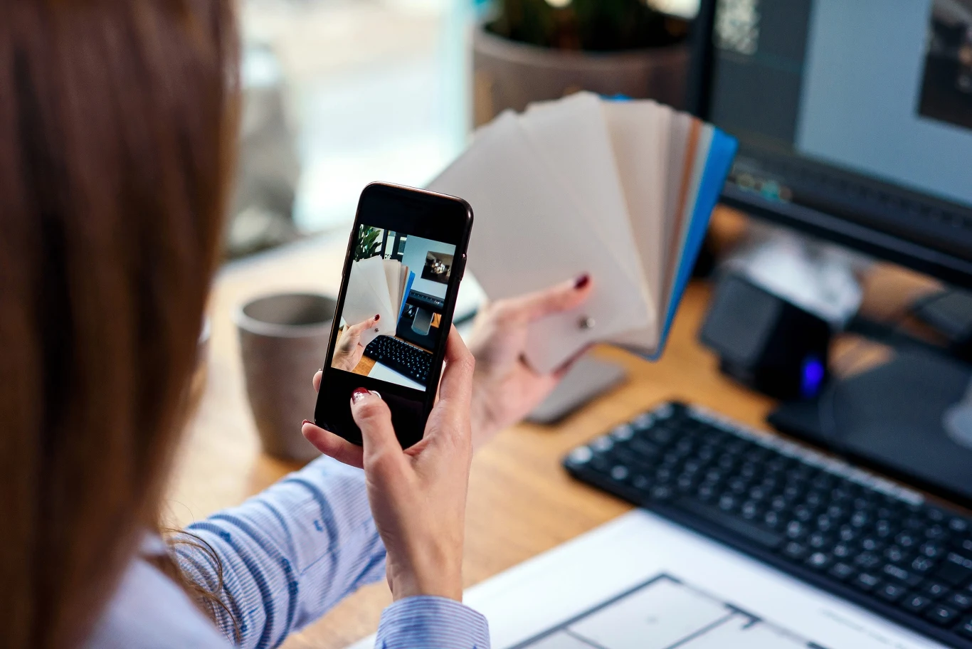Zapisujesz wszystko na telefonie? To może niszczyć twoją pamięć Osoba trzymająca telefon i wachlarz próbek kolorów fotografuje laptopa na biurku z dokumentami.