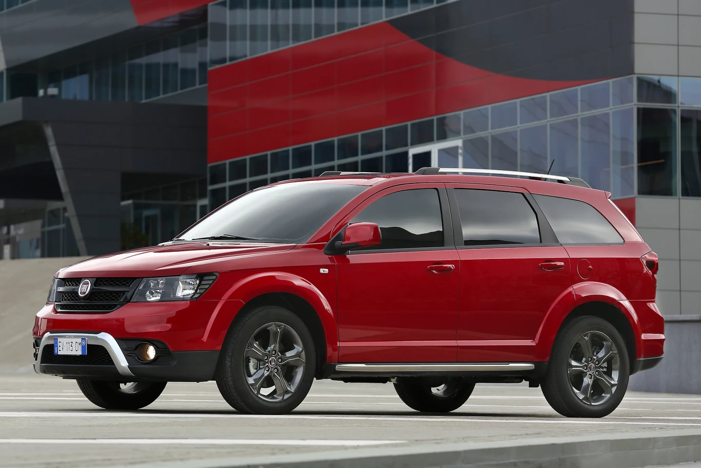 Fiat Freemont był wierną kopią Dodge'a. Otrzymał tylko inne logo i silniki Czerwony SUV Fiat Freemont ustawiony na tle nowoczesnego budynku z przeszklonymi i czerwonymi elementami fasady.