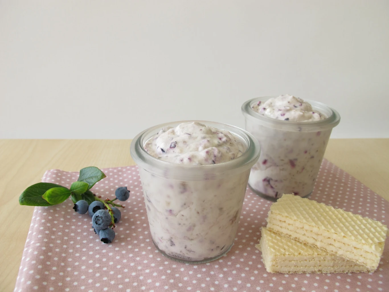 Najzdrowszy mrożony jogurt przygotujemy samodzielnie w domu Dwie szklanki kremowego deseru z jagodami, obok świeże jagody na gałązce i trzy wafle ułożone na różowej serwetce w białe kropki.