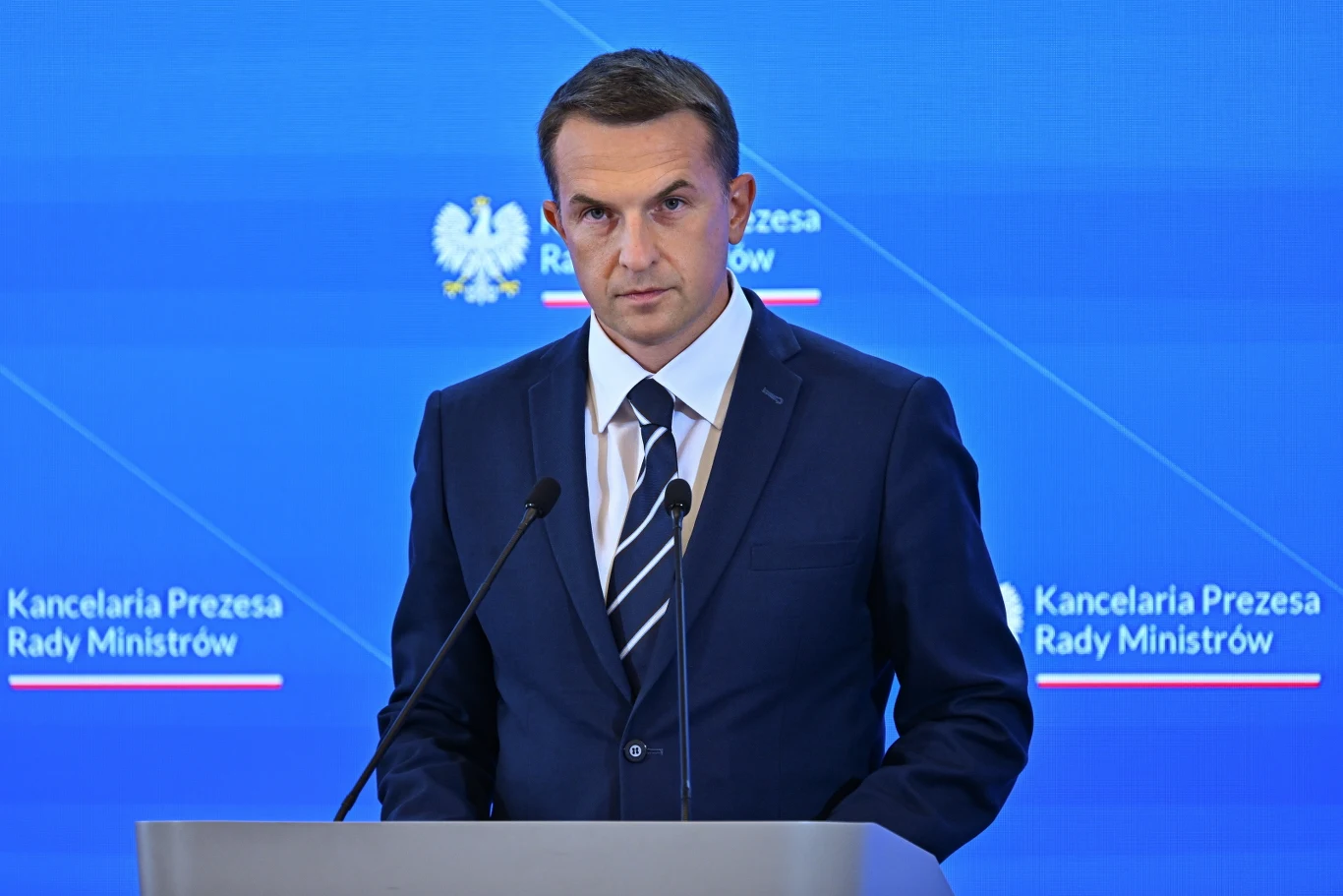 Rzecznik rządu Adam Szłapka Adam Szłapka za mównicą, w garniturze i krawacie, na tle niebieskiej ścianki z napisem Kancelaria Prezesa Rady Ministrów.