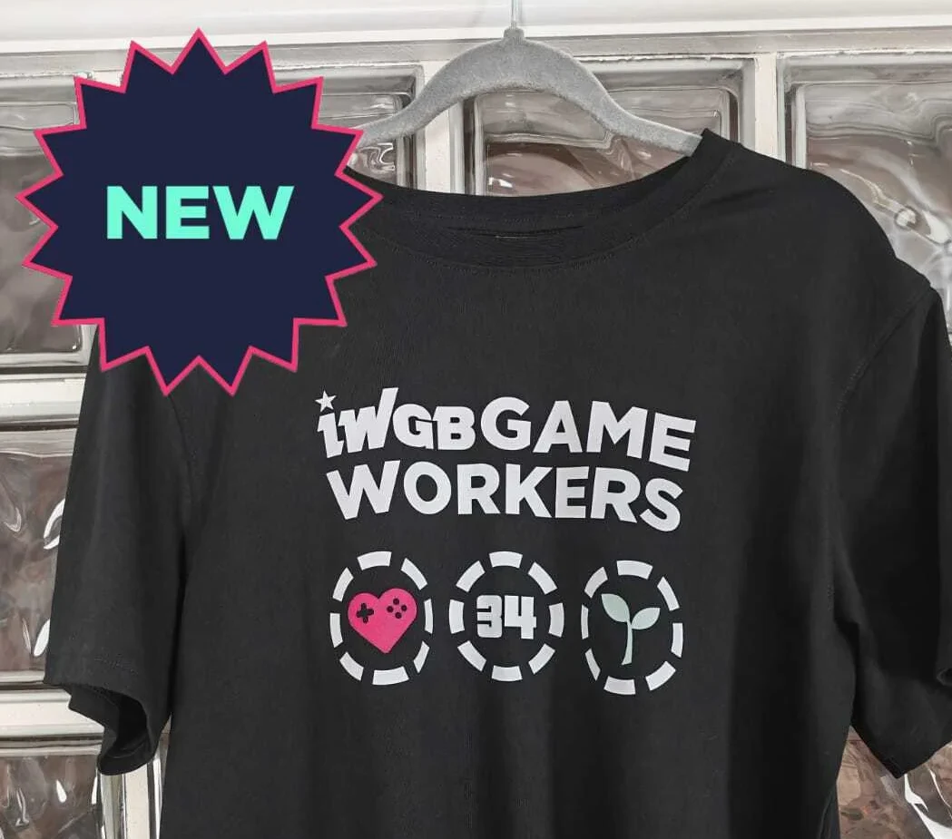 fragment zdjęcia limitowanej koszulki Czarna koszulka z białym napisem 'IWGB GAME WORKERS' oraz trzema symbolami: sercem z kontrolerem do gier, liczbą 34 i sadzonką w okrągłych ikonach, zawieszona na szarym wieszaku na tle szklanych pustaków.