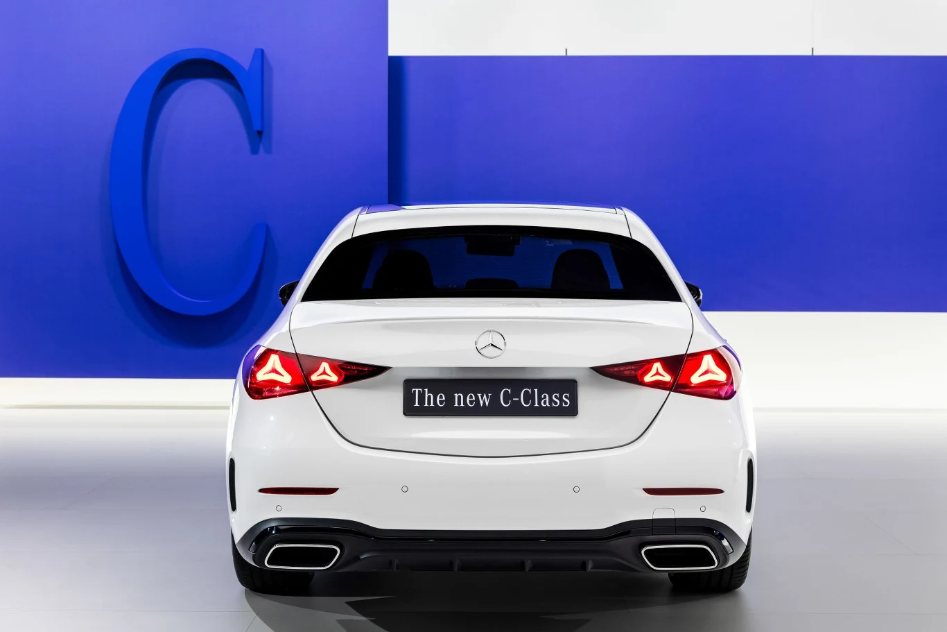 Ceny Mercedesa klasy C zaczynają się od 199 900 zł. Biały Mercedes-Benz C-Class zaparkowany na tle ściany w kolorze niebieskim z dużą literą C, widok od tyłu podkreśla nowoczesny design tylnych świateł i logo marki.