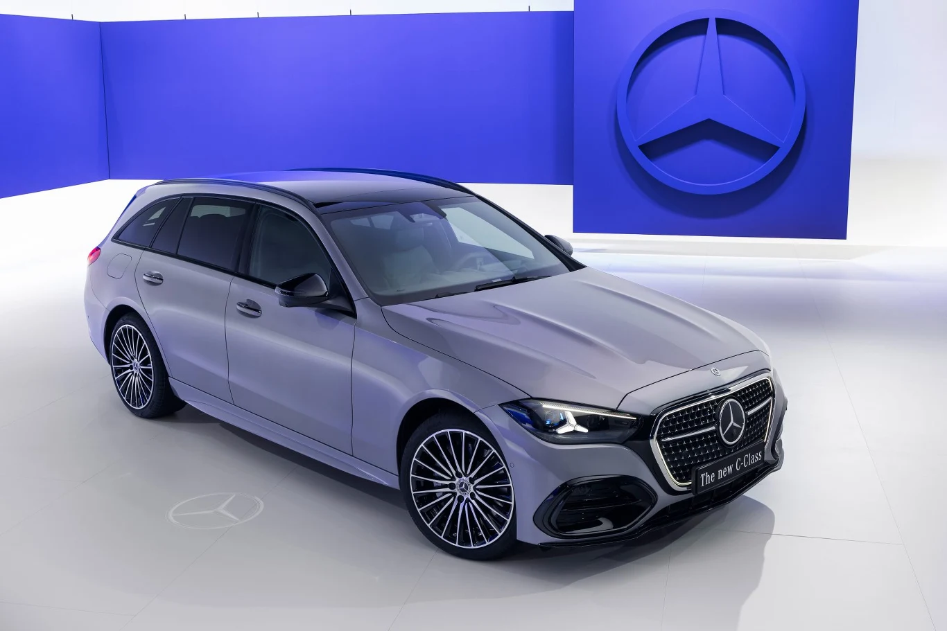 Po modernizacji Mercedes klasy C dostępny jest w wariantach benzynowych, wysokoprężnych i hybrydowych typu plug-in. Srebrny samochód Mercedes klasy C w wersji kombi na tle niebieskiej ściany z dużym logo marki Mercedes-Benz.
