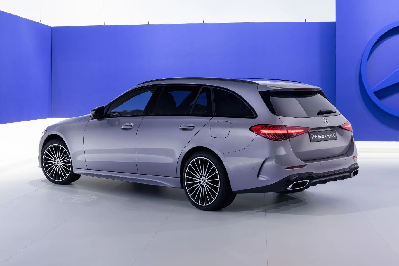 Mercedes klasy C zwraca na siebie uwagę m.in. sygnaturą świetlną nawiązującą do symbolu marki. Wyjątkiem są tylne światła w kombi. Nowoczesny samochód kombi marki Mercedes o opływowych kształtach, metaliczno-szare nadwozie, czarne sportowe felgi i charakterystyczne tylne światła LED na tle niebieskiej ściany z dużym logotypem marki.