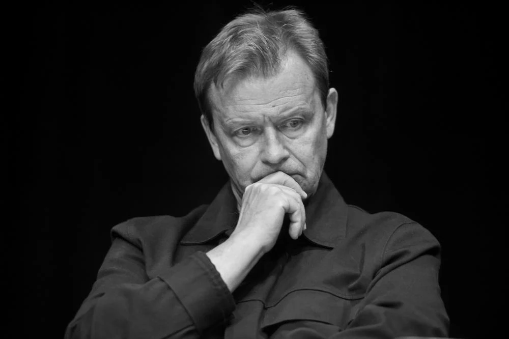 Jan Frycz Jan Frycz opiera głowę na dłoni, ubrany w ciemną kurtkę, czarne tło.