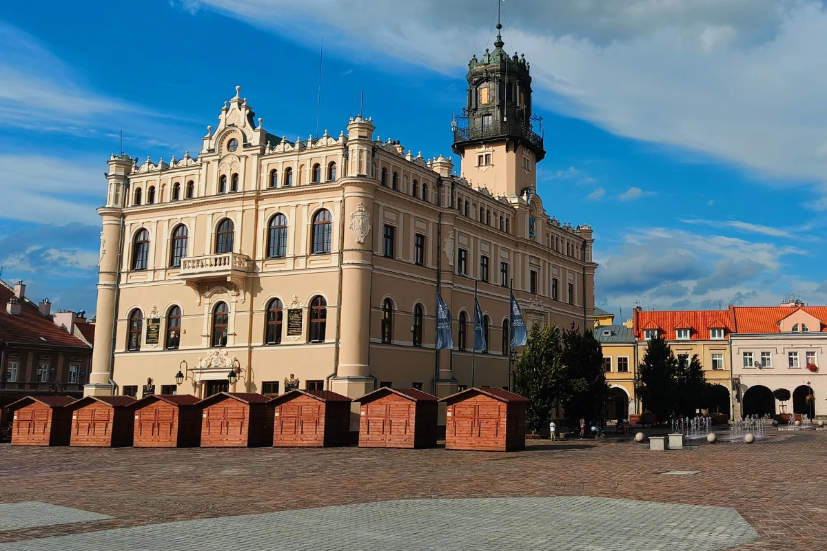 Zabytkowy budynek ratusza z wysoką wieżą i bogatą fasadą, otoczony szeregiem drewnianych domków jarmarcznych ustawionych na brukowanym rynku.